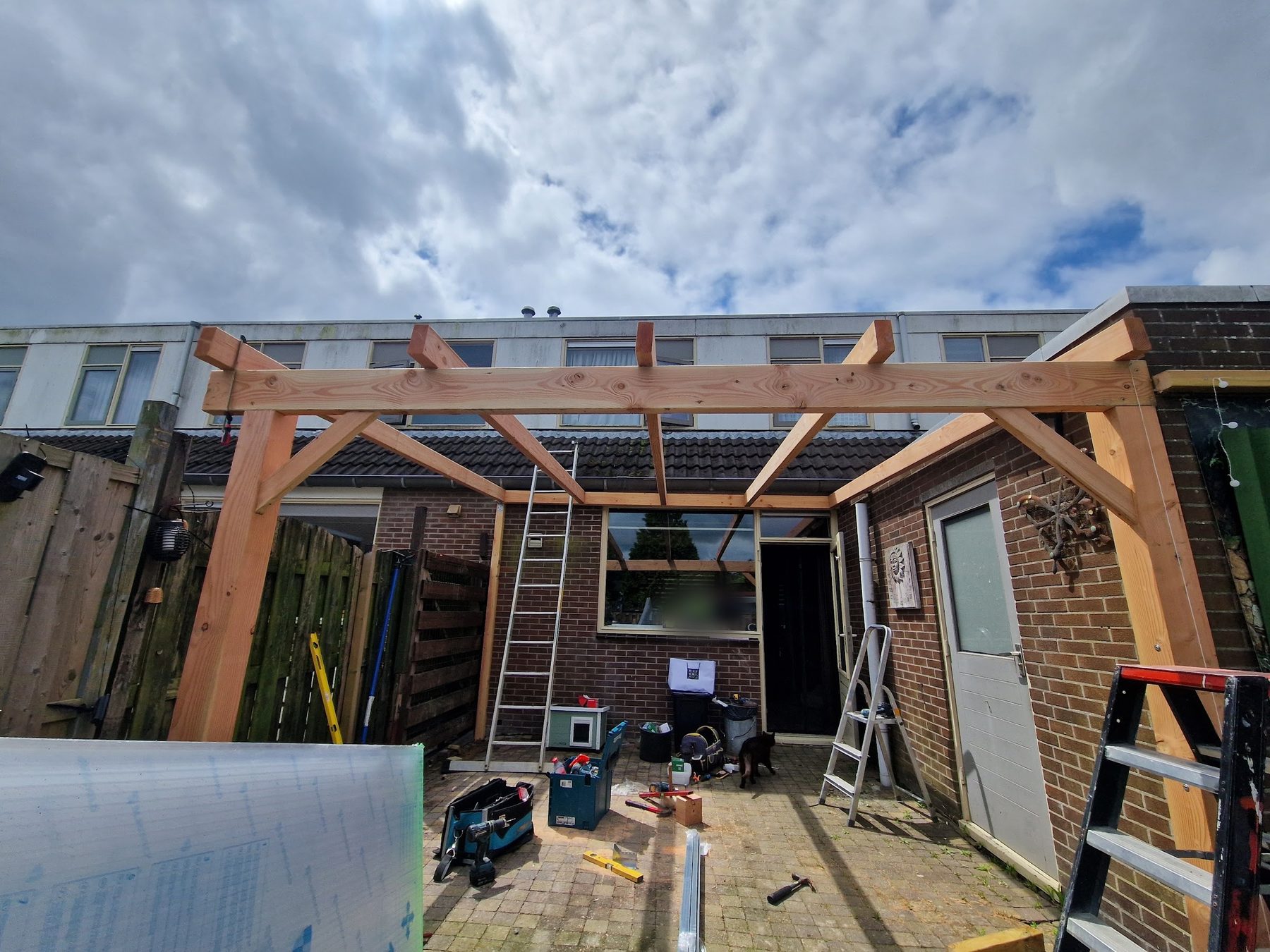 Een Douglas houten overkapping in aanbouw tegen de achtergevel van een rijtjeshuis, met ladders en gereedschap op het terras